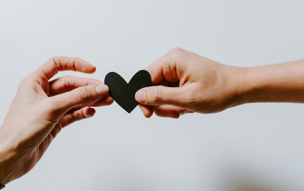 Two hands reaching in to hold a small black paper heart. Symbolising relationships and potentially other concepts related to love.